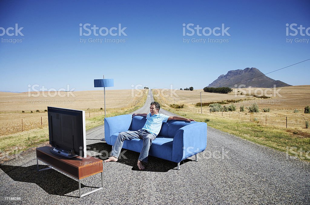 Man watching television on his sofa, smoking a pipe, under a lamp. 